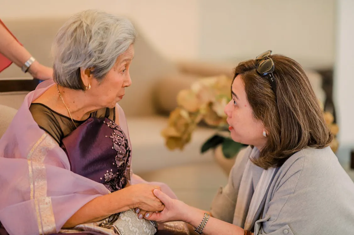 Adult daughter leaning in with gentle concern while holding her elderly mother's hands