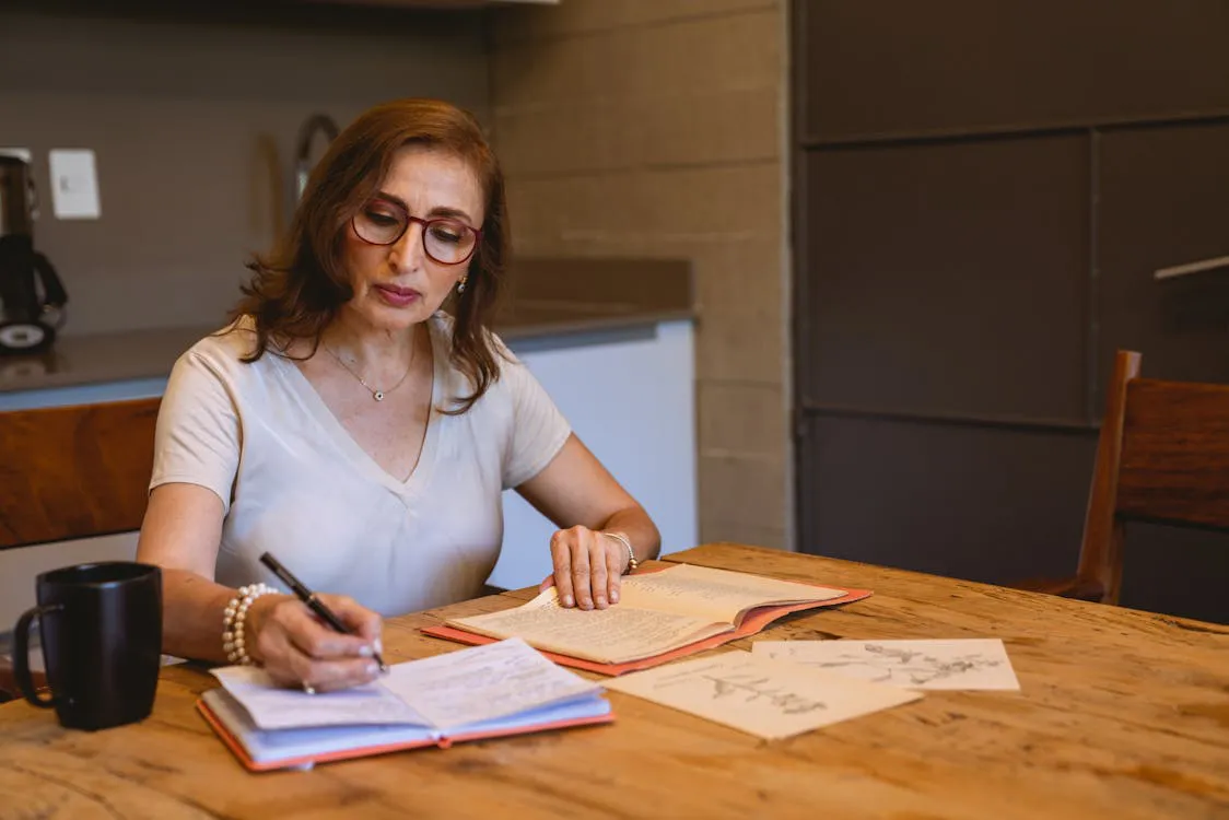 Middle-aged woman writing careful notes in a notebook at a warm kitchen table