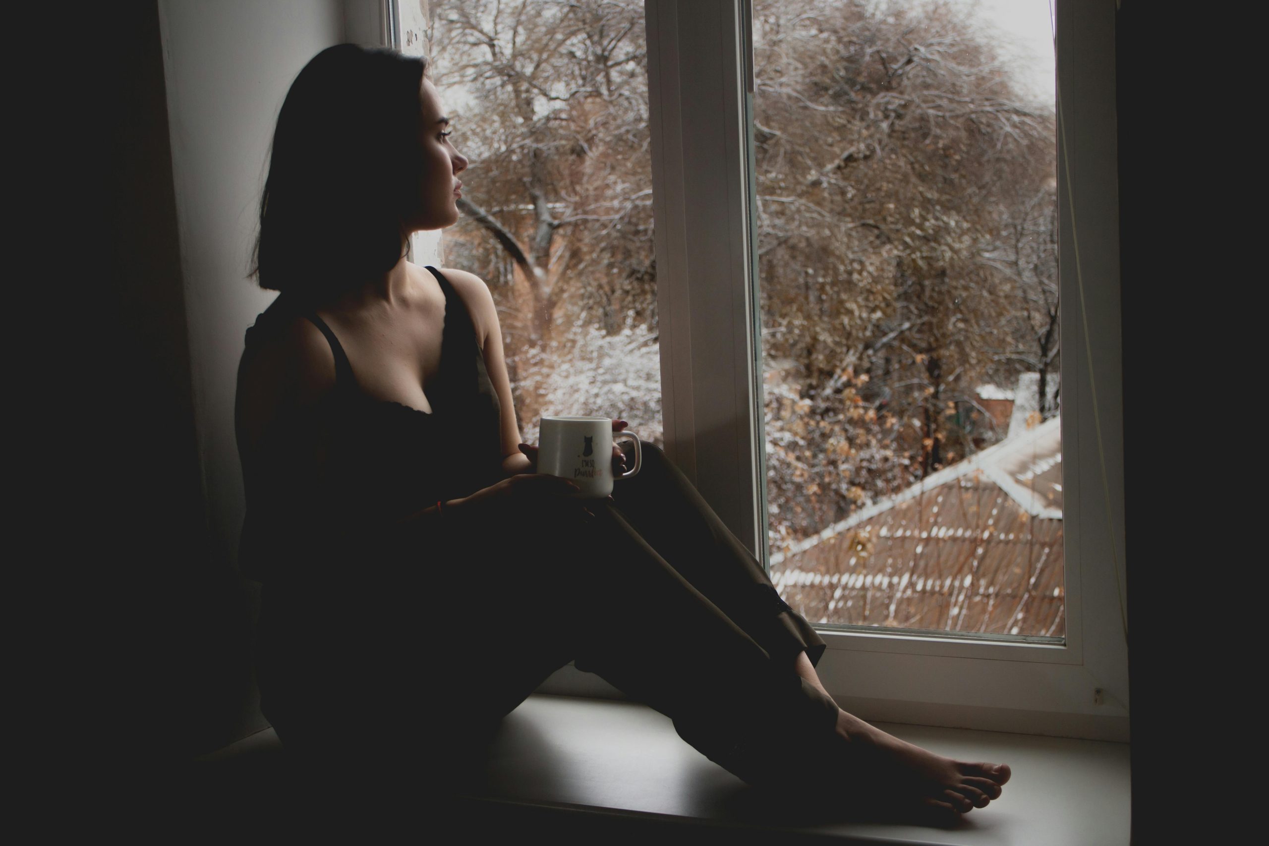 Woman sitting by a window in the late afternoon, holding a mug and looking outside thoughtfully
