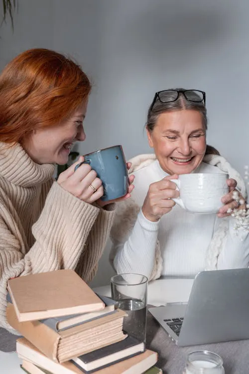 Adult daughter and mother laughing together over warm coffee mugs at home