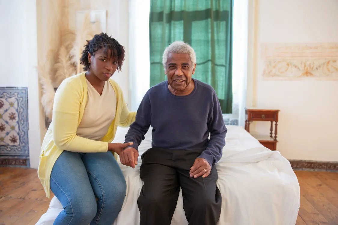 Adult daughter sitting beside elderly father, holding his hand with calm, caring presence in a home bedroom