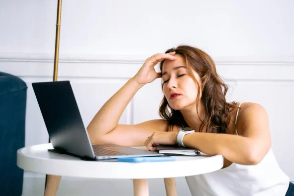 Exhausted middle-aged woman with eyes closed, hand on forehead, resting at a small table