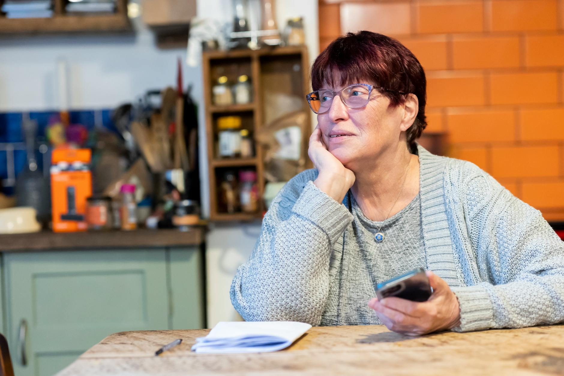 Caregiver at kitchen table in the evening, phone in hand, reviewing notes by lamp light