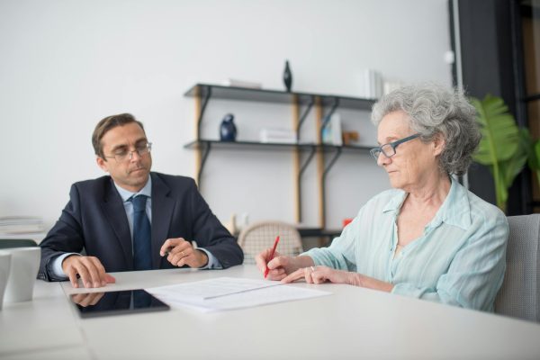 Elderly woman signing paperwork at a desk with a consultant reviewing documents together